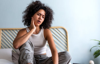 young woman with curly hair touching cheek in pain having toothache sitting on bed