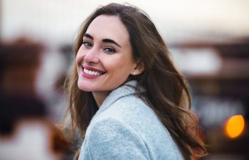 A woman with long brown hair and a light gray coat smiles at the camera outdoors, with a blurred background.